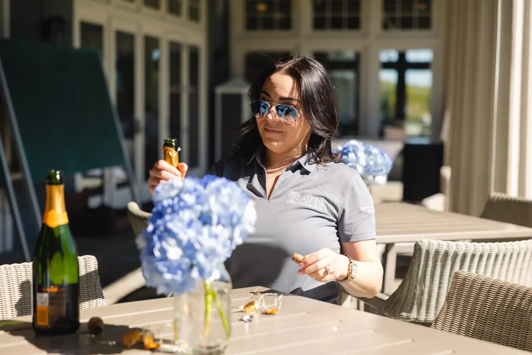 Volunteer in sunglasses arranging blue hydrangeas on a clubhouse patio table beside a champagne bottle