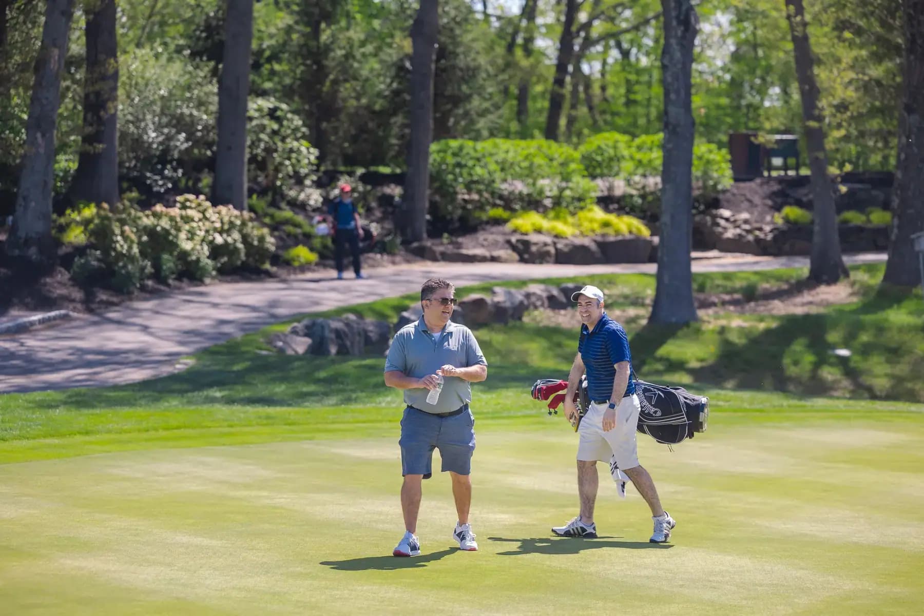 Two golfers with a bag walking across a green below a wooded hillside