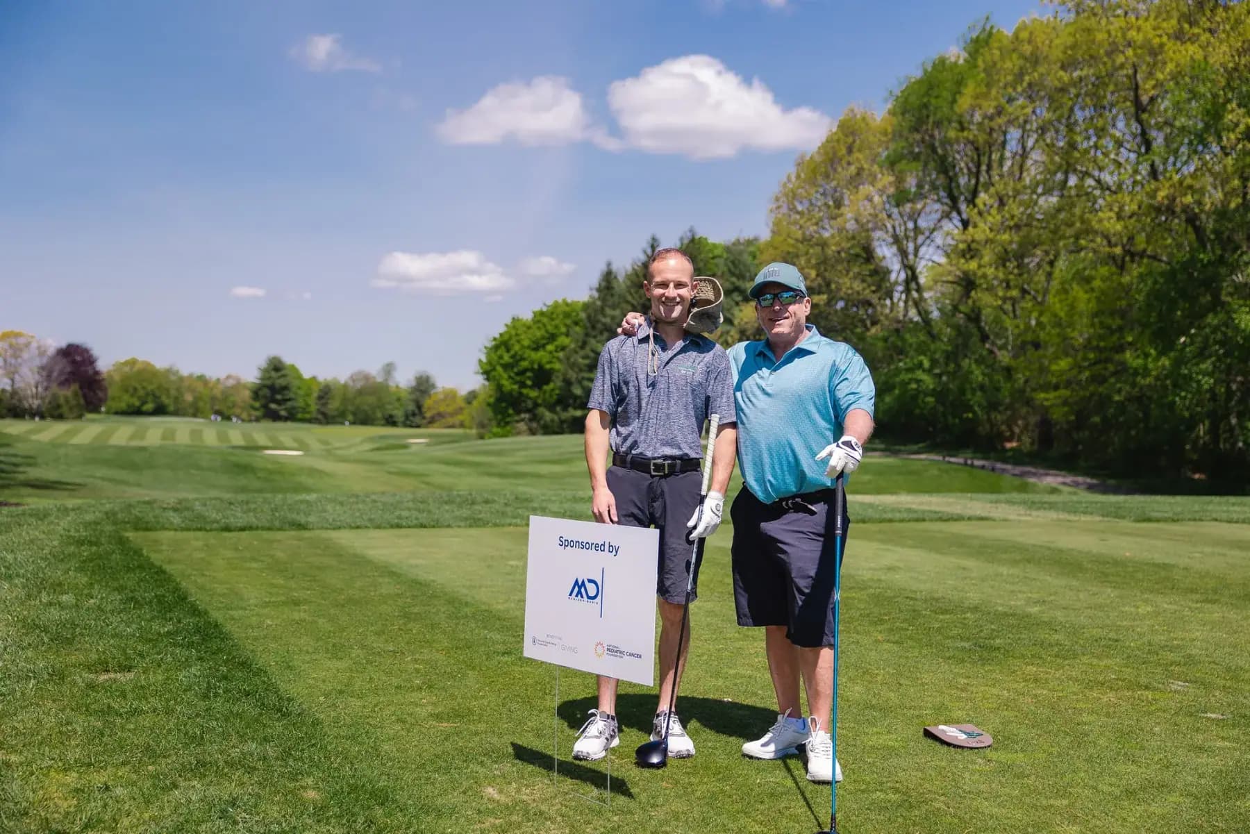 Two golfers posing beside a hole sponsor sign on a fairway under blue sky