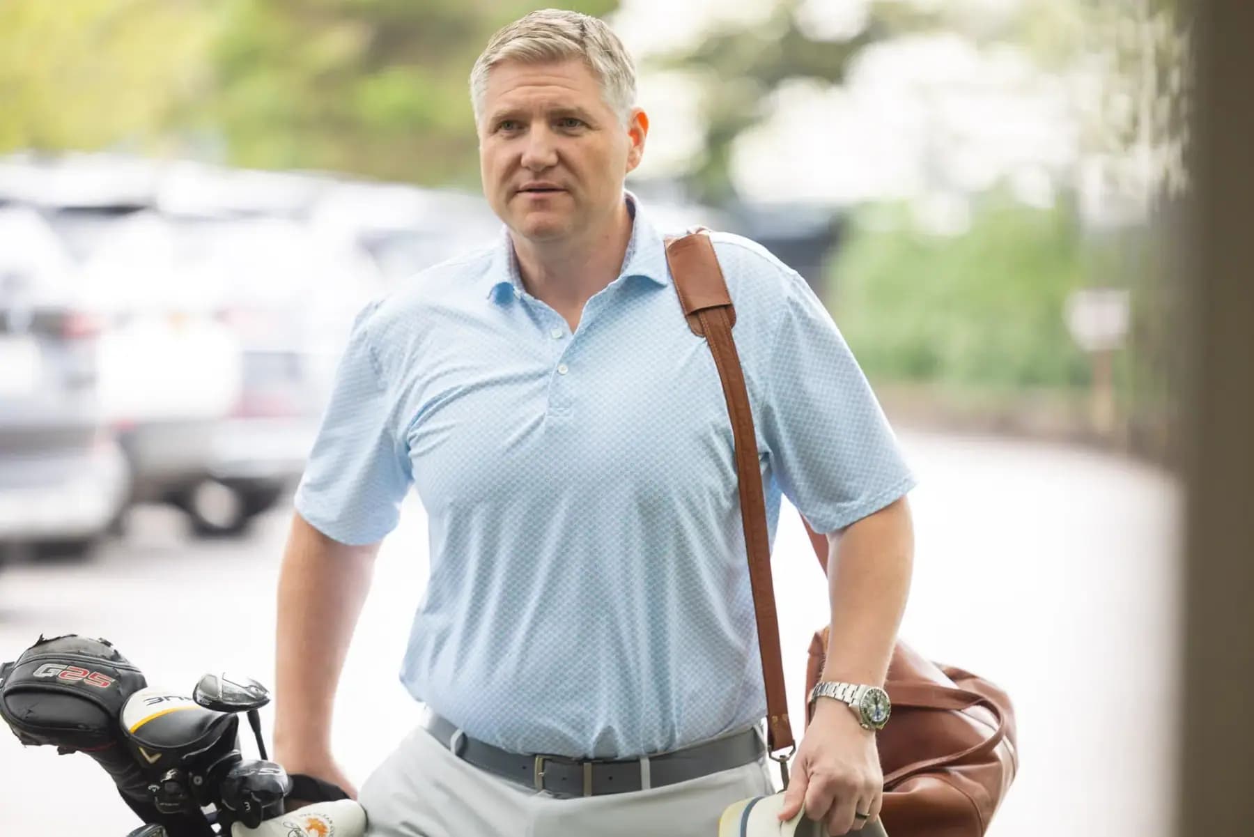 Golfer in a light blue polo carrying a leather bag past parked golf carts