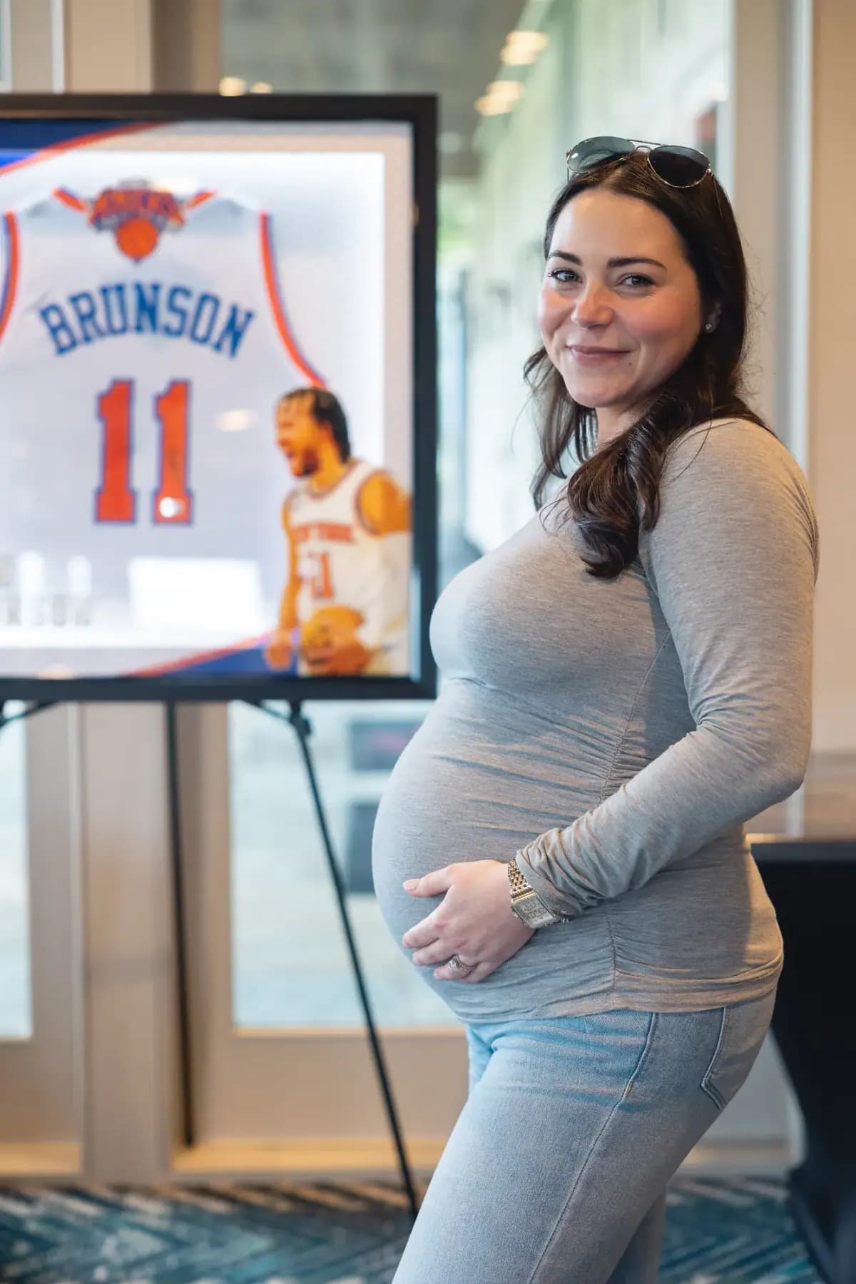 Smiling pregnant woman beside a framed signed Knicks Brunson jersey