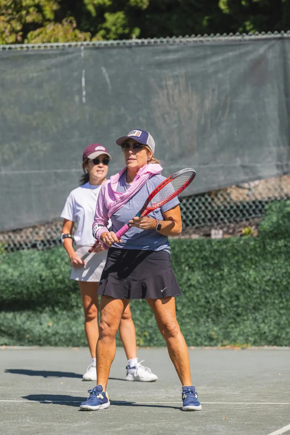 Player in a pink scarf and black skirt holding a racket on court, another player behind