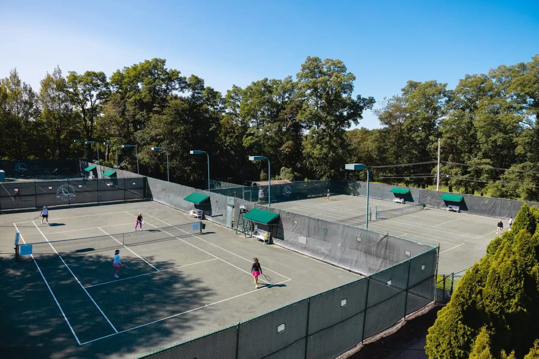 High view over several clay tennis courts surrounded by trees under blue sky