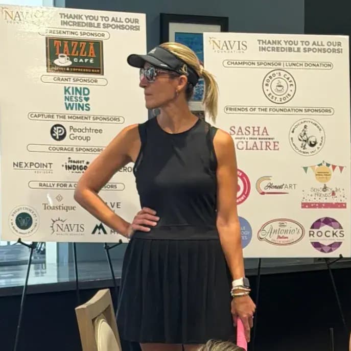 Woman in a black tennis dress and visor standing by the sponsor thank-you boards
