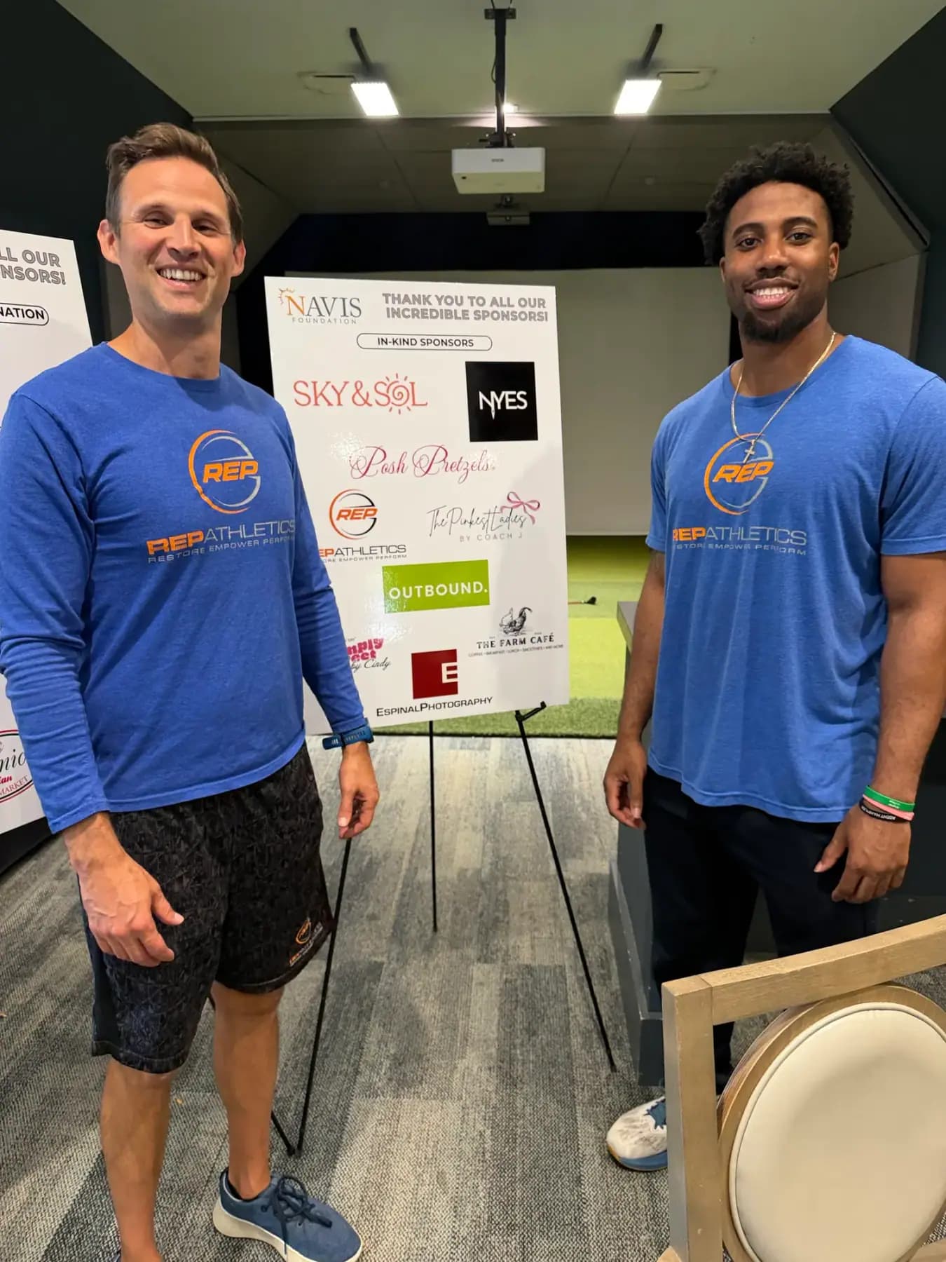 Two men in blue shirts posing beside the sponsor boards