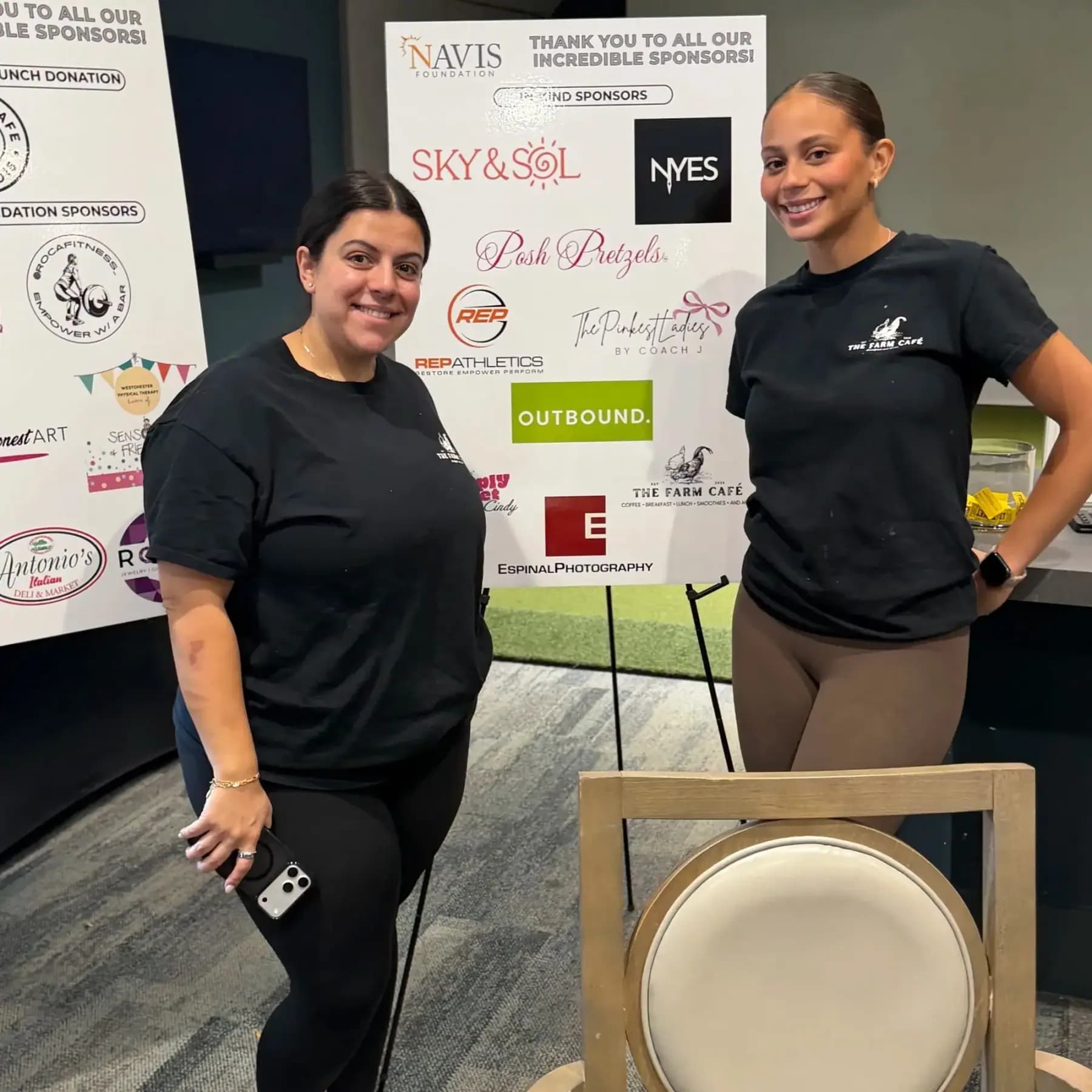 Two women in black T-shirts posing beside the sponsor boards